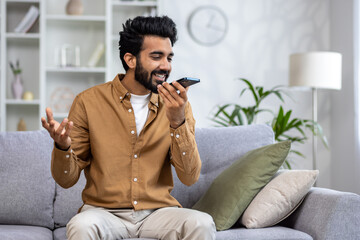 Joyful man speaking into smartphone like a microphone on a cozy sofa, expressing happiness and...