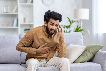 Worried man sitting on sofa, hand on head, expressing stress or headache in a well-lit living room with minimalist decor.
