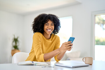 Happy smiling young African American woman sitting at home table and looking at camera while holding mobile phone using smartphone shopping online in app on cellphone in living room. Portrait.