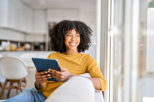 Happy African American lady using tab device looking away on sofa at home. Smiling pretty young woman sitting on couch relaxing looking away at window holding digital table in kitchen.