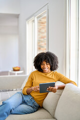 Happy young African American woman sitting on couch at home using digital tablet. Smiling relaxed...