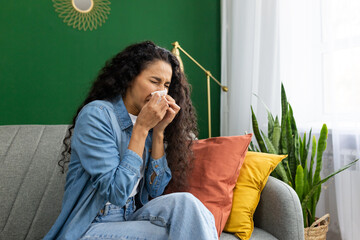 Woman in denim shirt feeling unwell, sneezing into tissue on couch, home interior with plants,...