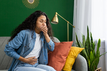 A distressed young woman feeling nauseous on a couch, hand over her mouth, trying to suppress the...