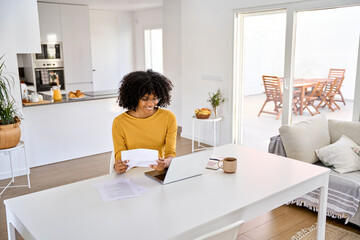 Fototapeta na wymiar Young smiling happy African American woman checking financial paper calculating banking payments using laptop computer paying bills online sitting at home table in modern kitchen interior.