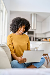 Happy young African American woman sitting on couch at home using laptop. Smiling ethnic lady relaxing on sofa looking at computer technology device browsing web doing online shopping in living room.