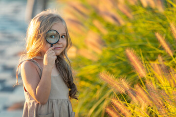 Fototapeta na wymiar Cute adorable Caucasian girl looking at plants grass in park through magnifying glass. Child looking through a magnifying glass on the grass in summer day. Child education, early development. concept.