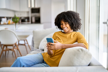Happy smiling young African American woman customer sitting at home on sofa looking at cellphone...