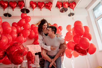 Playful, romantic moment between crazy couple near red balloons in bright room with white interiors. Woman sitting on mans back. Couple fooling around, laughing, looking into each others eyes. Life