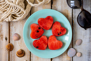 Summer flat lay. Watermelon hearts on a blue plate on wooden background. Sun glasses, shells, sea stones. Vacation mode, holiday inspiration, travelling concept