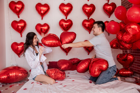 Playful moment between couple engaging in pillow balloons fight on bed, with heart shaped red balloons in background. Woman is joyfully fooling around with man. Bed is strewn with romantic rose petals