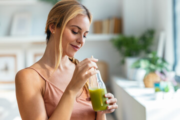 Kind woman drinking a healthy green smoothie standing in the living room at home.