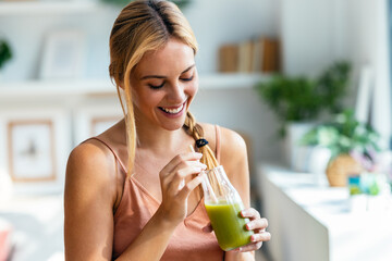 Happy woman drinking a healthy green smoothie standing in the living room at home.
