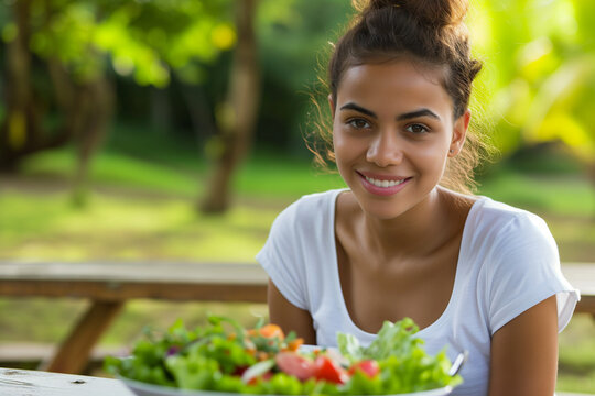 A smiling young woman is sitting at a wooden table outdoors, enjoying a colorful salad on a bright sunny day.
