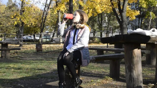 a beautiful girl sits at a table in an autumn park, headphones on her neck, a disposable cup with a warming drink in her hands. the child dangles his legs. hobbies, recreation and entertainment