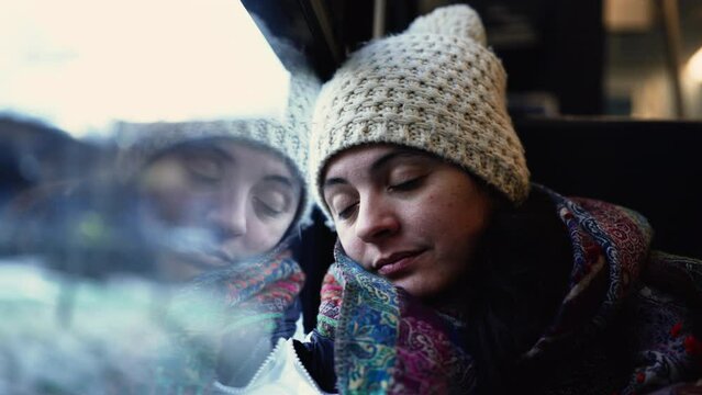 Passenger Asleep on Train with Moving Landscape View. Woman leans head on window traveling during winter season, wearing scarf and beanie