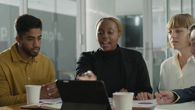 Four young adults in businesswear talking and using digital tablet during meeting in office