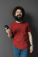 Joyful man in a red t-shirt and hat holding a smartphone, sharing a candid laugh against a dark background