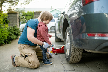 Cute toddler boy helping his father to change car wheels at their backyard. Father teaching his...