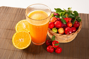 Fresh Organic Acerola and Orange Juice in a glass cup with sliced orange fruit and acerola berries in a bamboo basket in white background in top view