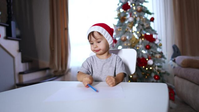 Caucasian baby in red Santa cap drawing with pencil. Little kid points at his picture with finger. Christmas tree at backdrop.