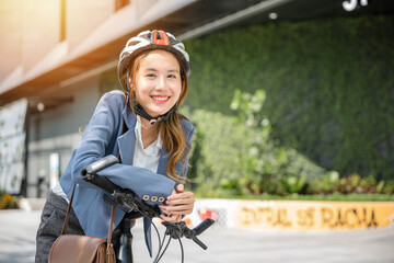In the city an Asian businesswoman helmeted and in a suit stands with her bicycle ready for a cheerful morning commute to the office. This image combines work and outdoor fun. - obrazy, fototapety, plakaty