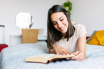 Happy young woman lying on bed while reading a book. Leisure activity concept.