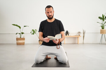 Caucasian guy with beard looking and reading book of mudras. Concentrated middle-aged man leaning...