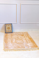 A muslim carpet set on the floor, beige brownish color. A closed Holy coran book placed on the rug...