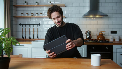 Young curly hair man playing video game on a tablet computer while sitting in the kitchen at home. Excited concentrated male engrossed in interesting game with tablet device	