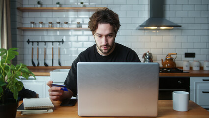 ,A curly hair man sitting in the kitchen entering credit card number on laptop for makes secure...