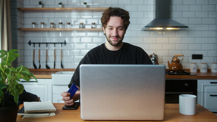 Smiling young curly hair man sitting in the kitchen entering credit card number on laptop for makes...