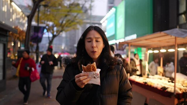 韓国のソウルの明洞の雪の降る寒い夜にナイトマーケットでスナックを買って食べるロングヘアーの30代の韓国人男性 A long-haired Korean man in his 30s who buys and eats snacks at the night market on a snowy cold night in Myeong-dong, Seoul, South Korea