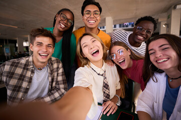 A large group of smiling and cheerful young multiracial university students taking a selfie with cell phone looking happy at camera. Generation z friends having fun together in the faculty classroom