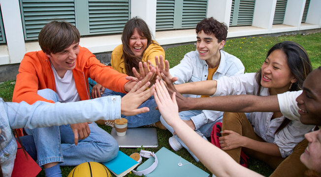 Laughing young people gathered in circle joining hands celebrating some academic success. Group multiracial students together sitting on grass outside high school building. Generation z in community.
