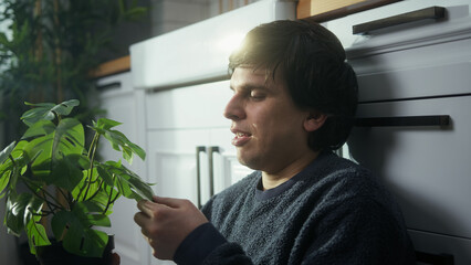 Young man sitting on the floor in the kitchen showing love and smells the flower in the pot he...