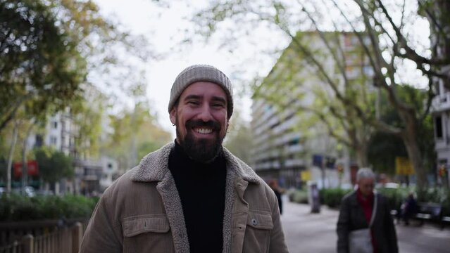 Handsome young man in winter clothes and hat walking outdoors looking camera. Smiling and confident male crossing a garden area in a city street. People on their way to a meeting