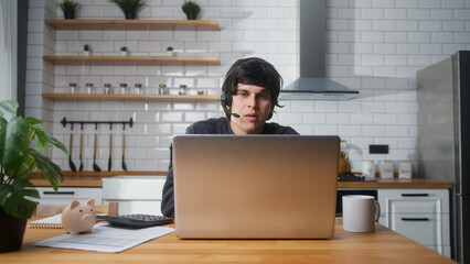 Young man sitting in kitchen at home wear wireless headset with microphone having video conference...