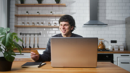 Smiling young man sitting in the kitchen entering credit card number on laptop for makes secure...