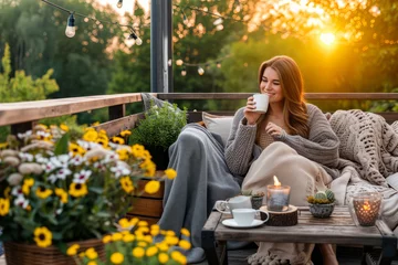 Fotobehang Young woman having a cup of tea on cozy wooden terrace with rustic wooden furniture, soft colorful pillows, light bulbs and flower pots. Charming sunny evening in summer garden. © MNStudio