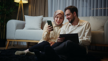 A married couple leaning against the sofa sitting on floor, woman using smartphone, husband using a...