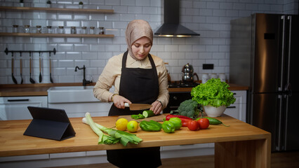 Vlogger young female chef with headscarf in an apron recording video cooking lesson about vegetarian meal or salad in modern kitchen at home. Online broadcast, woman leads food blog or course	