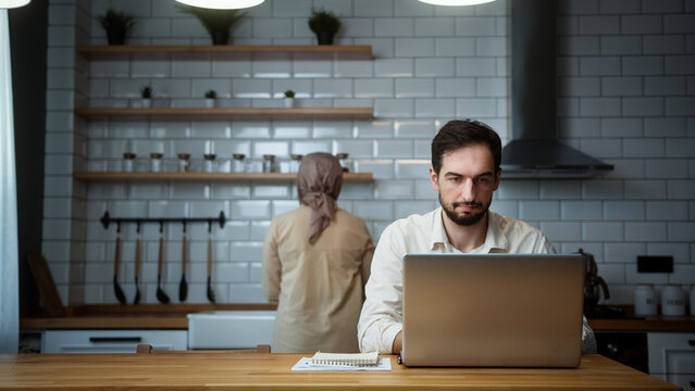 Young man is working on his laptop in the kitchen at home while his wife working in kitchen	
