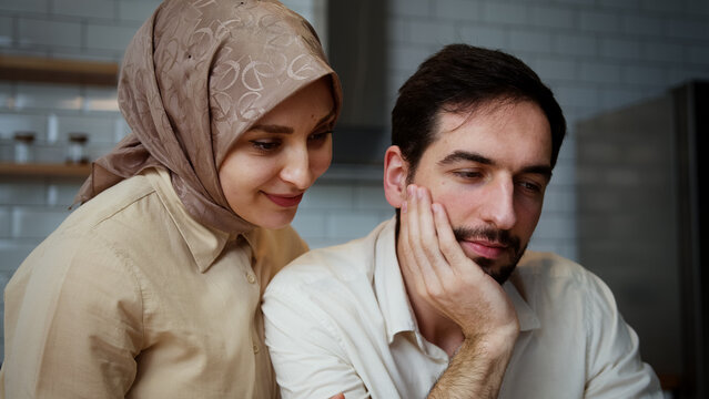Handsome man is working and typing on his laptop in the kitchen when his wife comes round from behind and hug	
