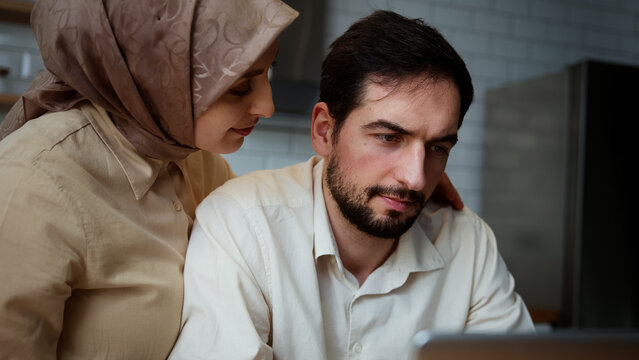 Close up of young man is working and typing on his laptop in the kitchen when his wife comes round from behind and hug	
