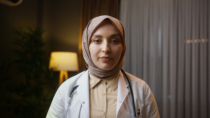 Portrait of female doctor in headscarf wears white coat looking at camera at night in home office