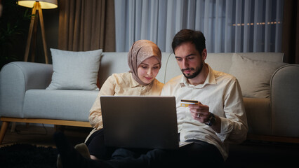 Young married couple leaning against the sofa sitting on floor at home at night, using a laptop to...