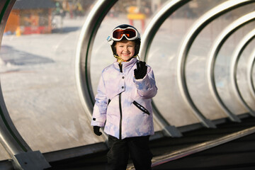 A girl on skis stands on a carpet ski lift in a glass tunel