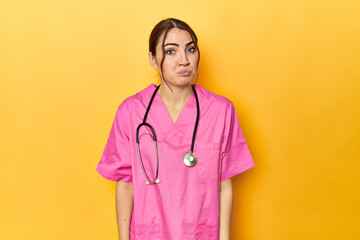 Nursing assistant in yellow background shrugs shoulders and open eyes confused.