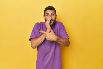 Young Hispanic man on yellow background saying a gossip, pointing to side reporting something.