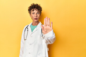 Caucasian mid-age female doctor on yellow studio standing with outstretched hand showing stop sign,...